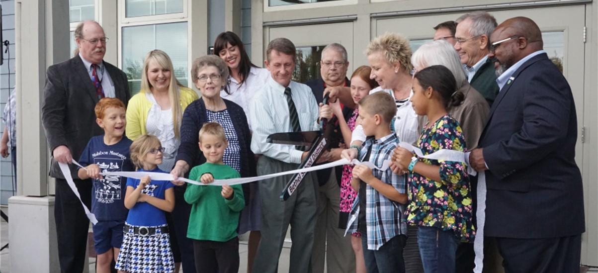 Group of people surrounding a man holding giant scissors cutting a ribbon in front of a building